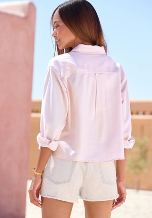 Woman with long brown hair wearing a light pink shirt with rolled sleeves and light denim shorts, standing outdoors in sunlight.