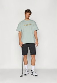 Svetlozelené oversize tričko s nápisom „Range Rats“ v žltej farbe, v kombinácii s čiernymi šortkami a bielou športovou obuvou na mužskom modeli.