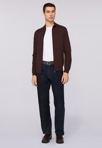 Bruine ritssluitingjas over een wit shirt, gecombineerd met donkere broek en zwarte schoenen. De outfit heeft een slank, modern ontwerp.
