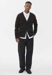 Brūns adīts kardigāns ar V veida kakla izgriezumu un sešiem pogām, valkāts uz balta krekla un kombinēts ar melnā krāsā biksēm un apaviem. Vienkāršs, ikdienišķs dizains.