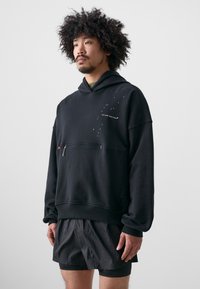 Svart huva sweatshirt med en framficka, som har tryckt text och stjärnmotiv. Bärs med svarta sportshorts; mjukt tyg.