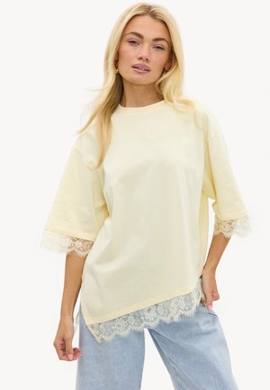 Mujer rubia con una camiseta grande de color amarillo pálido con detalles de encaje en las mangas y el dobladillo, combinada con vaqueros azul claro, posando con una mano en la cadera.