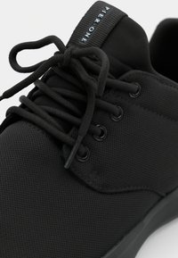 Zwarte sneaker met een bovenwerk van textuurstof, ronde veters, metalen oogjes en een merklabel aan de hiel met "PIER ONE" in het blauw.