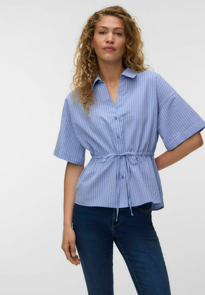 Femme aux cheveux bouclés portant une chemise bleu clair à manches courtes boutonnée avec des rayures verticales et une taille nouée, associée à un jean bleu foncé.