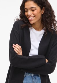 Μαύρη, υφασμάτινη ζακέτα με ανοιχτό μπροστινό μέρος, μακριά μανίκια και δύο τσέπες μπροστά, φορεμένη πάνω από λευκό T-shirt και ανοιχτόχρωμο τζιν.