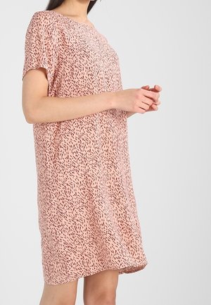 Vrouw draagt een loszittende, kortere mouw, knielange lichtroze jurk met een klein donker gespikkeld patroon op een effen achtergrond.