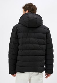 Sort polstret jakke med hætte, der har et quiltet design, glat overflade og ribstrikkede manchetter. Bagfra viser den detaljerne på midtersømmen.