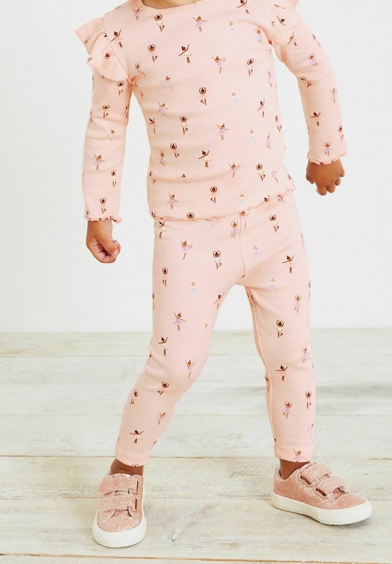 Tout-petit portant un haut à manches longues rose à volants et un legging avec un imprimé ballerine, associé à des baskets roses pailletées à scratchs, debout sur un sol en bois clair.
