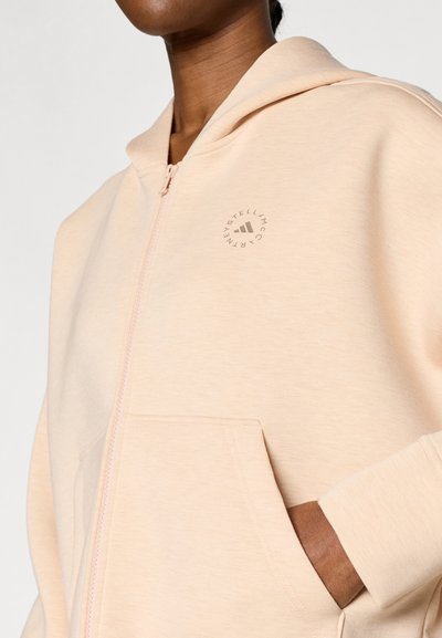 Ανοιχτό ροδακινί ζακέτα με φερμουάρ, σε απαλό ύφασμα, με μπροστινή τσέπη και κυκλικό λογότυπο στην αριστερή πλευρά. Η κουκούλα προσθέτει ζεστασιά και κάλυψη.