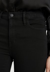 Black denim jeans featuring a high waist, front pockets, and a classic button closure with visible stitching details. Smooth texture.