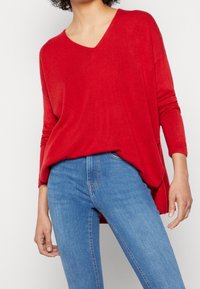 Suéter ligero de punto rojo con escote en V, mangas largas y un corte relajado, combinado con jeans de mezclilla azul claro.