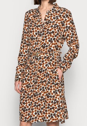 Kobieta ubrana w sukienkę na długi rękaw, zapinaną na guziki, w wzór kwiatowy w kolorach brązowym, czarnym i białym, z pasującym materiałowym paskiem, ręką wsuniętą do kieszeni.