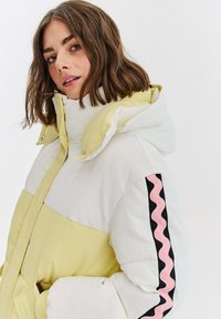 Bunda s váčkom v bielej a svetlo žltej farbe s rebrovanou textúrou. Obsahuje kapucňu a ružové vlnité akcenty na čiernom rukáve. Predné zapínanie na zips.