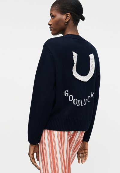 Γυναίκα με σκούτερο πουλόβερ που έχει ένα λευκό πέταλο και το κείμενο "GOOD LUCK" στην πλάτη, συνδυασμένο με παντελόνι με κόκκινες και λευκές ρίγες.