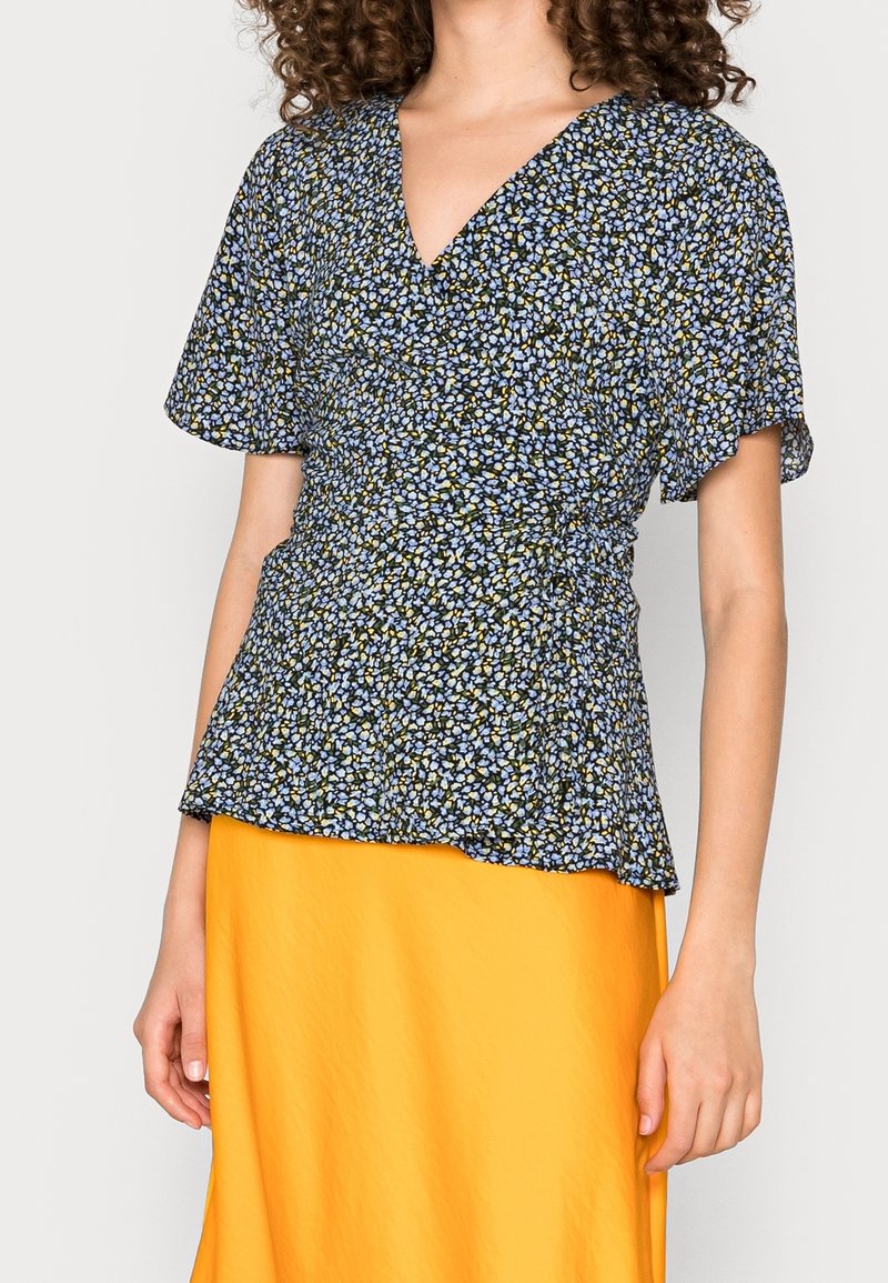 Femme portant un chemisier cache-cœur à manches courtes bleu à motif floral avec un décolleté en V profond et une jupe jaune vif.