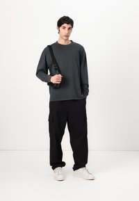 Long-sleeve dark grey shirt, black cargo pants, white sneakers, and a black shoulder strap, standing against a plain background.