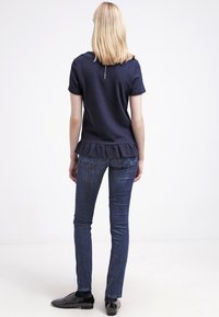 Haut à manches courtes en marine avec un ourlet à volants, associé à un jean en denim bleu foncé. La vue de dos montre un détail de fermeture éclair au niveau du cou et une coupe ajustée.