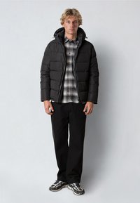 Sort pufferjakke med lodret syning og hætte, båret over en ternet flannel skjorte. Sorte bukser og mønstrede sneakers fuldender outfittet.