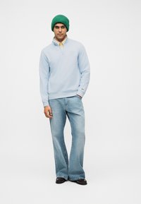 Homme portant un bonnet vert, un sweat-shirt à fermeture éclair partielle bleu clair, une chemise jaune en dessous, un jean bleu clair ample et des chaussures marron foncé, debout sur un fond blanc.