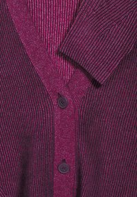 Gros plan sur un cardigan en maille côtelée violet et magenta avec deux boutons foncés et un tissu qui se chevauche au niveau du col et de la manche.