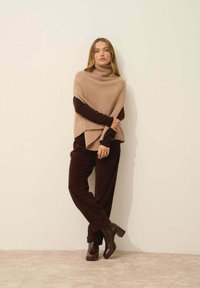 Beige žebrovaný rolák pončo přes hnědou dlouhou rukávovou košili, v kombinaci s tmavě hnědými kordovými kalhotami a robustními hnědými botami na podpatku.
