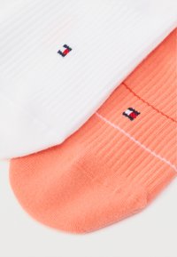 Δύο ζευγάρια κάλτσες έως τον αστράγαλο: οι λευκές με ριμπ υφή και λογότυπο, οι κοραλλί με απλή υφή και αντίθετο λογότυπο.