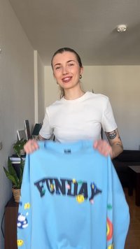 Νεαρή γυναίκα με τατουάζ χαμογελά ενώ κρατά ένα γαλάζιο φούτερ με τη λέξη «FUNDAY» και πολύχρωμα γραφικά με χαμογελαστά πρόσωπα και λουλούδια.