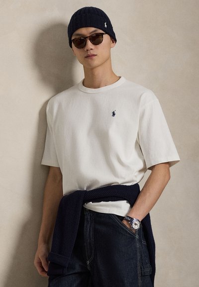 T-shirt blanc en coton à manches courtes avec un logo bleu marine, associé à un pantalon en denim foncé. Un bonnet en maille bleu marine et des lunettes de soleil complètent le look.