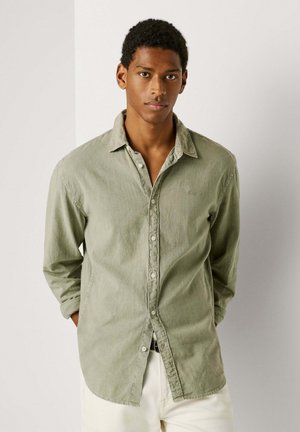 Junger Mann mit kurzem, lockigem Haar, der ein geöffnetes hellolivgrünes Hemd und weiße Hosen trägt, steht vor einer schlichten Wand.