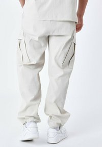 Lys beige cargo bukser med en tekstureret stof, der har sidelommer, elastiske cuffs og en afslappet pasform. Båret med hvide sneakers.