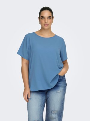 Kvinde med mørkt hår bundet tilbage, iført gyldne øreringe, en løs blå top med korte ærmer og slidte blå jeans med den ene hånd i lommen.