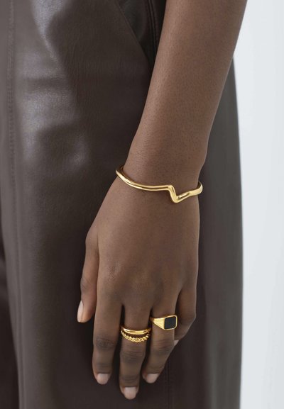 Kuldne käevõru lainekujundiga, virnastatud rõngad, millel on ruudukujuline must kivi ja tekstuurilised aktsendid, pruuni nahast taustal.