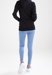 Person som bär en svart hoodie, ljusblå tajta jeans och vita Nike Air-sneakers, som går bortåt mot en vit bakgrund.