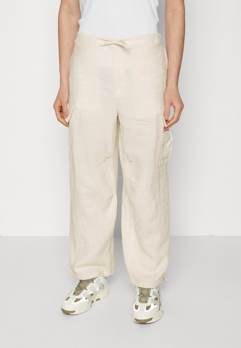 Pantalon cargo beige à jambes larges avec poches latérales, taille à cordon, porté avec des baskets blanches et vert olive et un haut blanc.