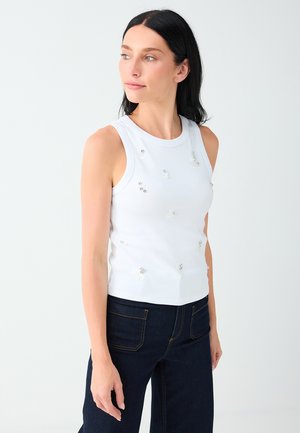 Femme aux cheveux foncés portant un haut blanc sans manches avec de petites décorations en perles et un jean bleu foncé, regardant de côté sur un fond uni.