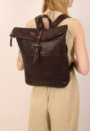 Femme portant une tenue sans manches beige avec un sac à dos en cuir marron foncé doté d'une fermeture éclair supérieure et d'une sangle à boucle, vue de dos.