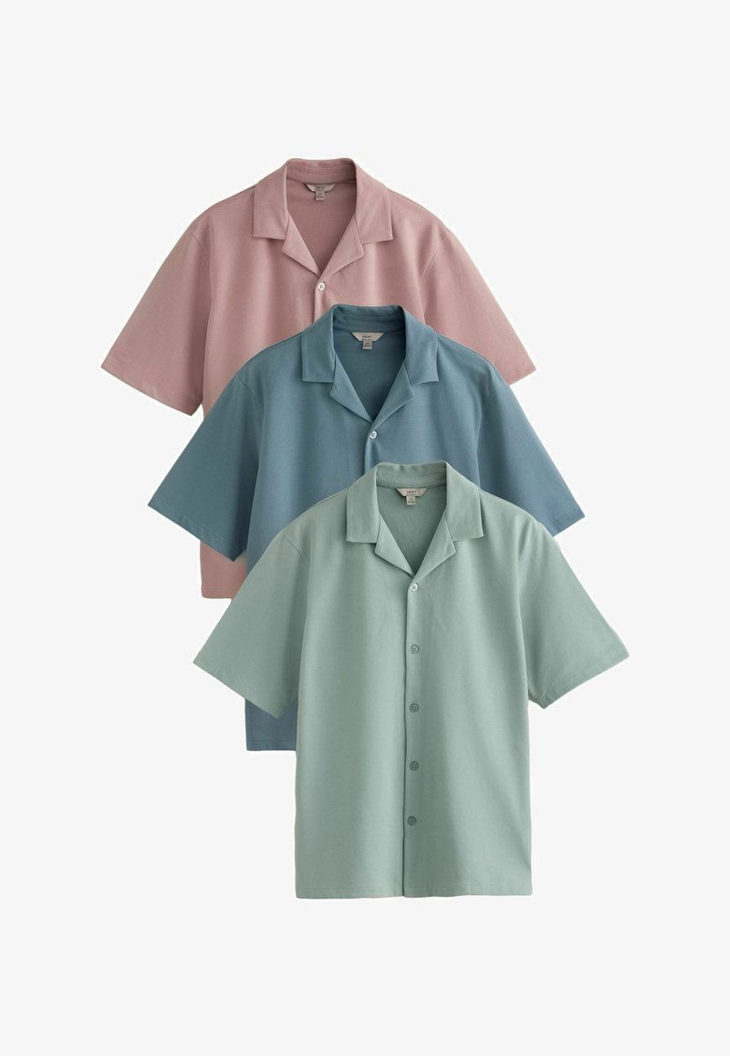 Trois chemises à manches courtes boutonnées avec col, en rose pastel, bleu et vert, disposées en chevauchement sur un fond blanc.