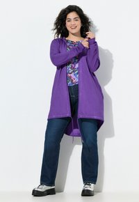 Langer lila Kapuzencardigan aus leichtem Stoff, mit offenem Design. Getragen über einem bunten Blumenoberteil, kombiniert mit dunkelblauen Jeans und silbernen Schuhen.