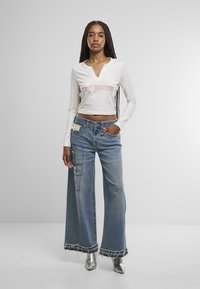 Bijela dugoruka cropped majica s natpisom "TRUE REASON", uparena s plavim širokim trapericama s patchwork detaljima i poderanim rubovima. Srebrne gležnjače.
