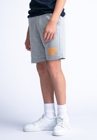 Shorts en coton gris avec poches latérales, ornés d'un logo imprimé orange sur la jambe gauche. Associés à des baskets blanches et des chaussettes.