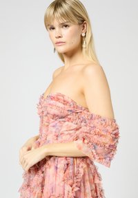 Olkaimeton mekko pehmeässä vaaleanpunaisessa kankaassa, jossa on kukkakuosi, ja jossa on röyhelöhihoja ja -reunoja. Mallilla on päällään suuret helmihyllykorvakorut.