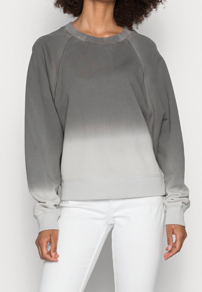 Personne portant un sweat-shirt à manches longues avec un dégradé gris-blanc et un pantalon blanc, debout devant un fond clair uni.