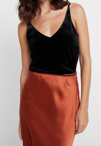 Camisole in velluto nero con un design a V, abbinato a una gonna in satin liscia di colore ruggine. L'outfit è caratterizzato da spalline sottili.