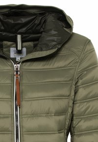 Veste matelassée vert olive avec doublure intérieure noire, couture horizontale, tirette de fermeture éclair en cuir marron et capuche rabattue.