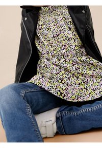 Persona che indossa un vestito floreale con fiori rosa, gialli e neri, sovrapposto a una giacca di pelle nera e jeans blu, seduta con le gambe incrociate.