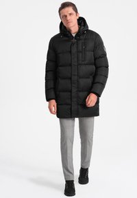 Cappotto imbottito nero con cappuccio, con chiusura a zip, tasche laterali e un design leggero trapuntato. Indossato con pantaloni grigi a quadri e stivali neri.