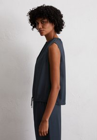 Frau mit lockigem dunklem Haar, die ein ärmelloses dunkelblaues Oberteil und passende Hosen trägt, steht seitlich gegen eine schlichte weiße Wand.