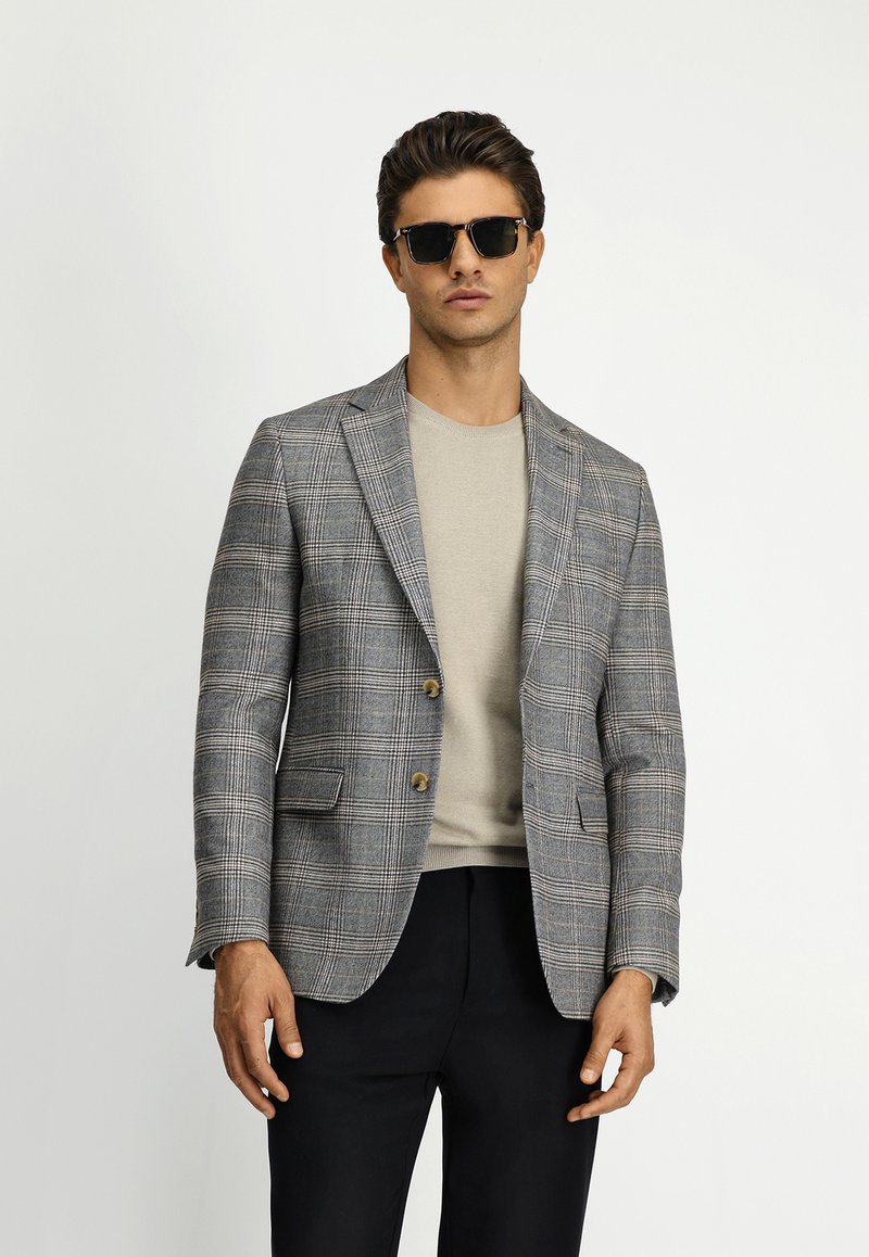 Grijs geruite blazer met twee knopen, notch revers en zijzakken, gedragen over een beige ronde hals T-shirt, gecombineerd met donkere broeken.