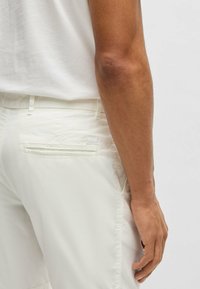 White pants with a textured finish, featuring a back pocket and stitched accents. A hand is positioned near the waistband.
