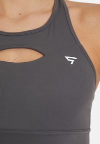 Siv ženski športni kratki top z izrezom, ki ima gladko teksturo in bel logo. Oblikovanje vključuje stil z racerback zadnjem delom.